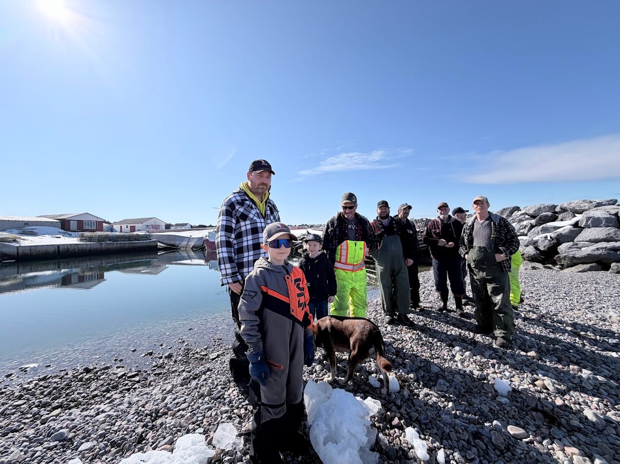 A group of men, children and a dog stand on a rocky beach.