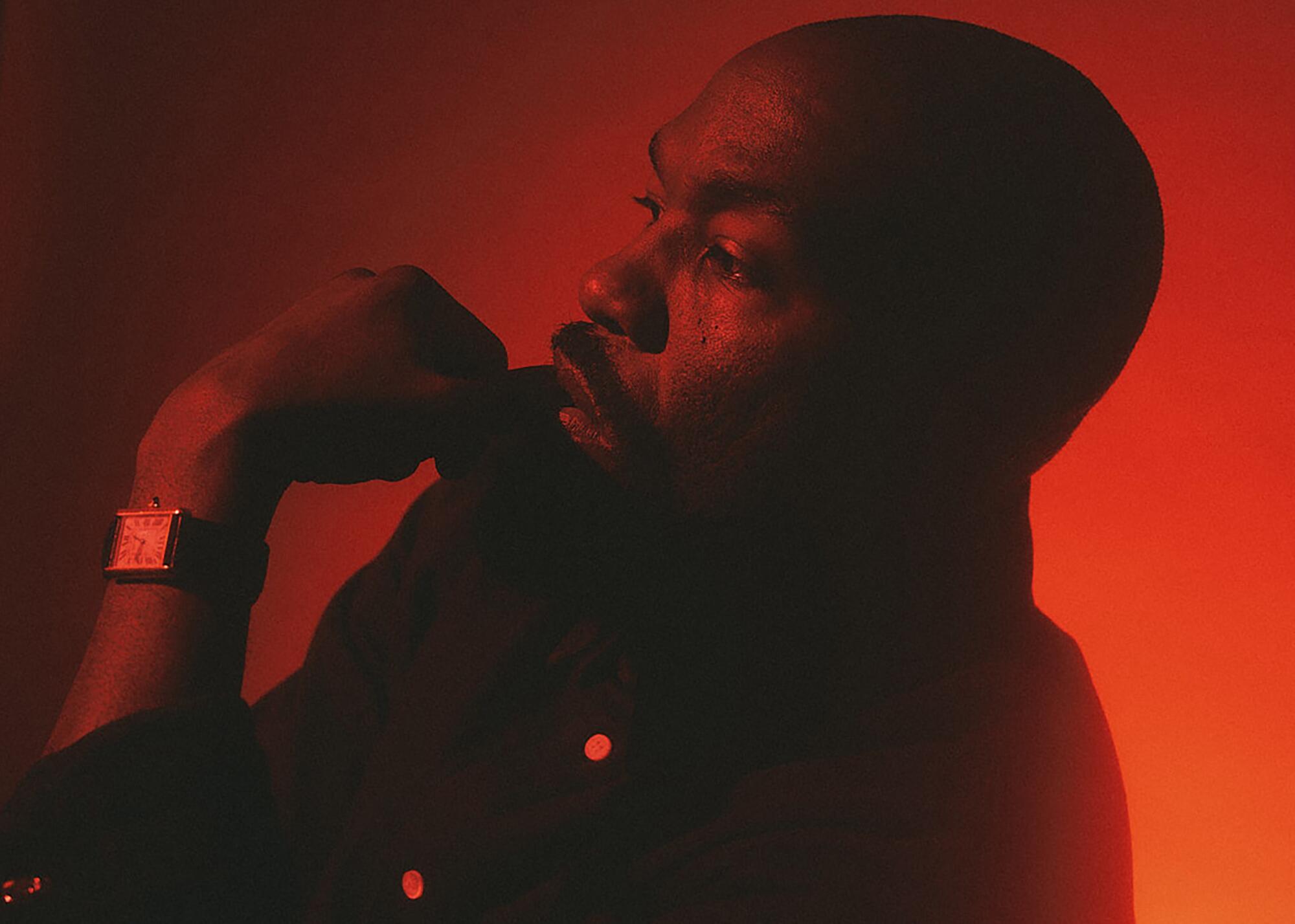 A man leaning back with his hand near his mouth in a red-lit room.
