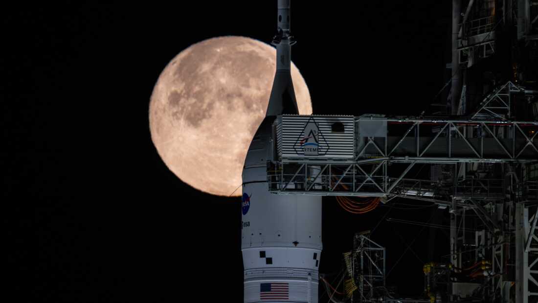 A large orange full moon rises behind a tall white rocket on a launch pad, with metal scaffolding around it at night.