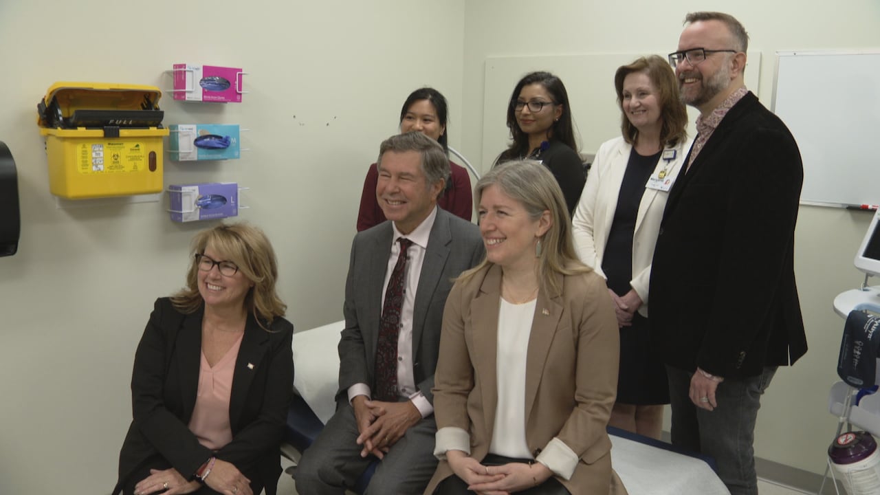 A group of six smiling people sit on a bed in an examining room in a doctor's office.