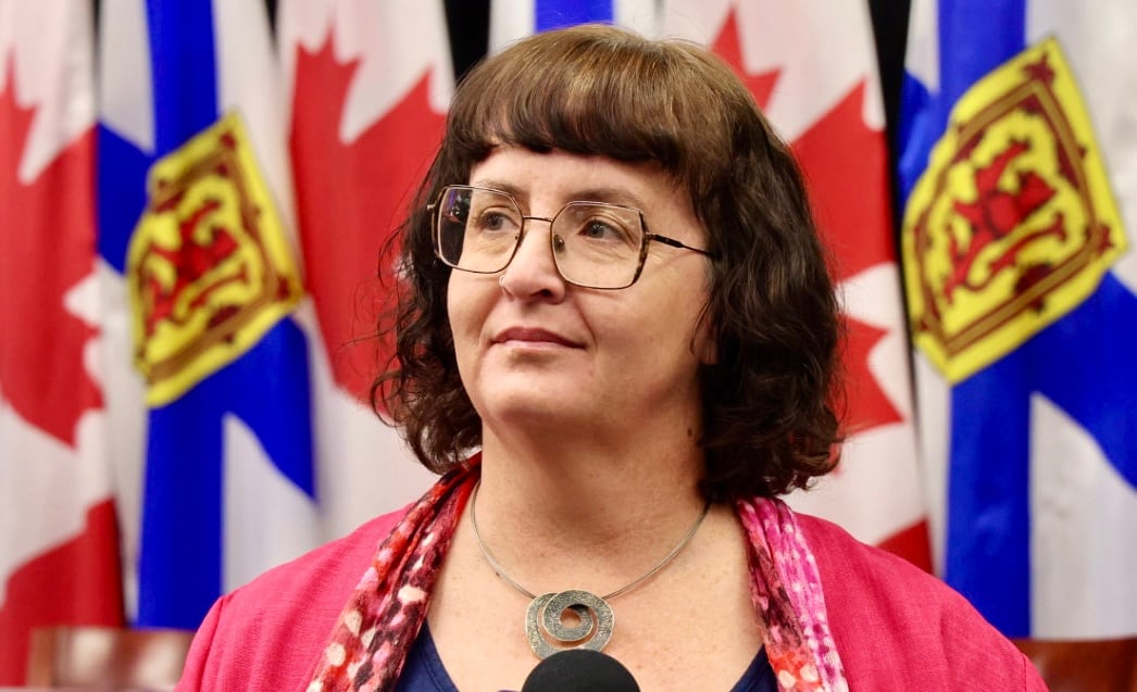 A person with glasses stands in front of Nova Scotia and Canadian flags.