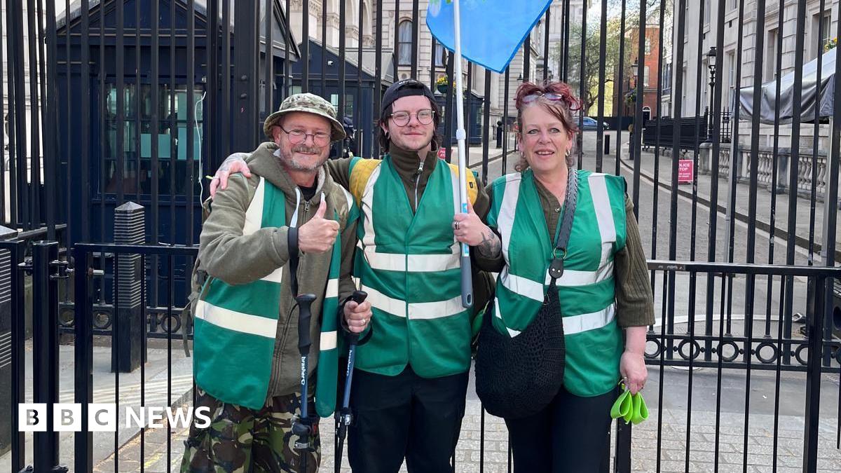 Rob, Lisa and son Drew outside gates in Downing Street. They are wearing dark green jackets and Rob Collins on the left is putting his thumb up.