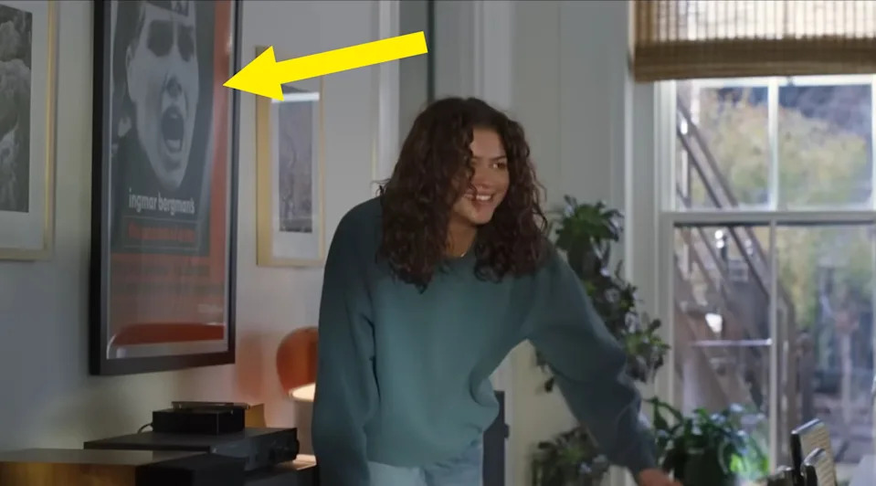 Person with curly hair in a casual outfit smiling in a living room with framed art on the wall