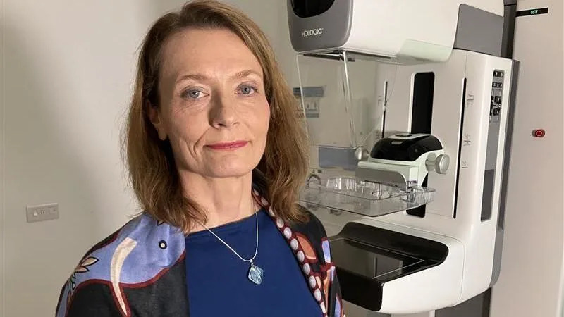 Dr Olga Strukowska, Consultant Radiologist and Director of West Sussex Screening service. Dr Strukowska is stood in front of the screening machine. She is wearing a patterned shawl and navy top. She has long brown hair.