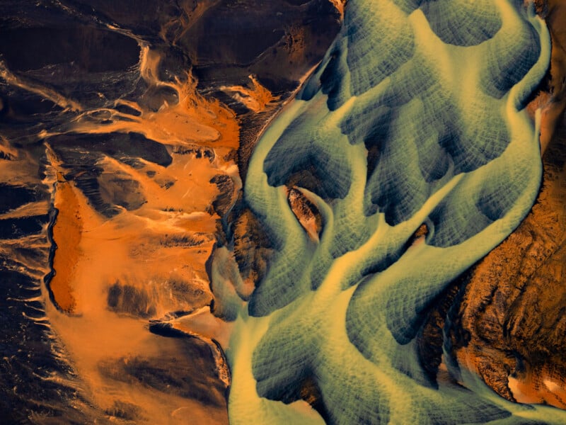 A surreal aerial view of a landscape with bright orange-brown terrain and winding blue-green river channels, creating an abstract, painterly pattern.