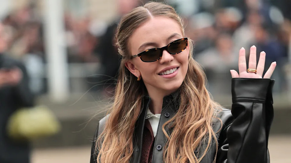 Sydney Sweeney with long wavy hair and sunglasses waves, wearing a black leather jacket, smiling.