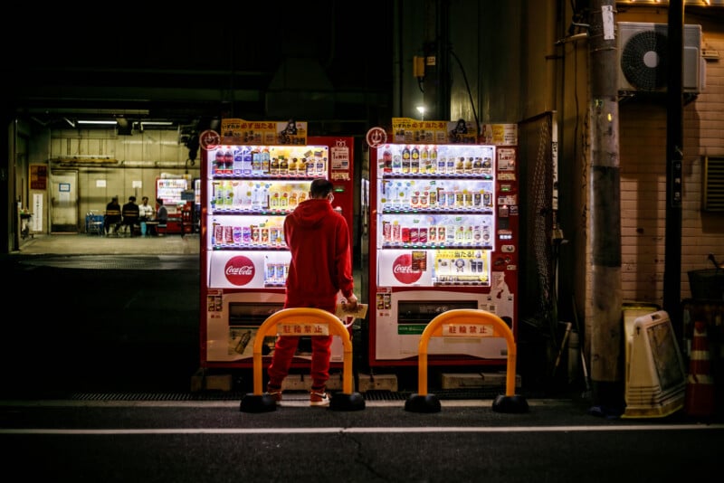 A person dressed in red stands at night in front of two brightly lit vending machines filled with drinks, located on a dark street with barriers in front of the machines.