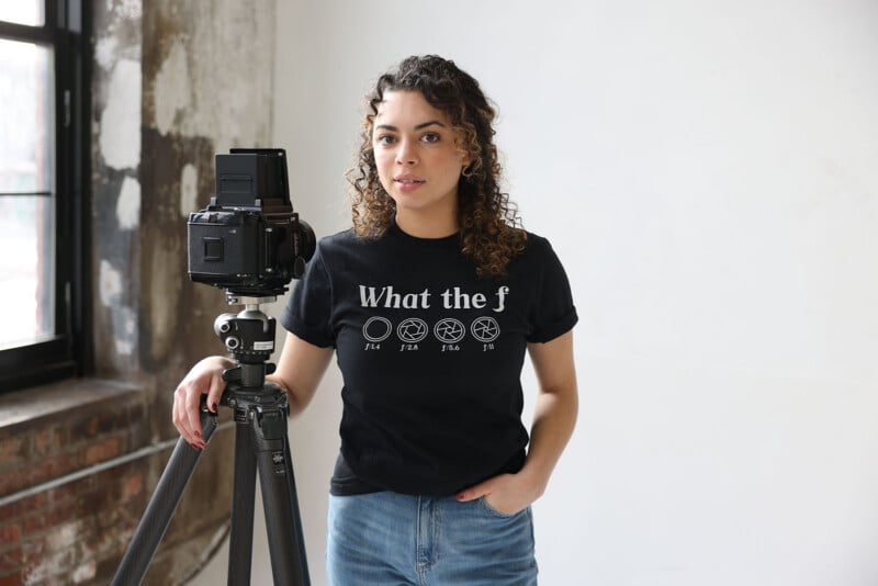 A woman with curly hair stands next to a camera on a tripod, wearing a black T-shirt that says "What the f" with drawings of camera aperture icons. She is indoors near a large window.