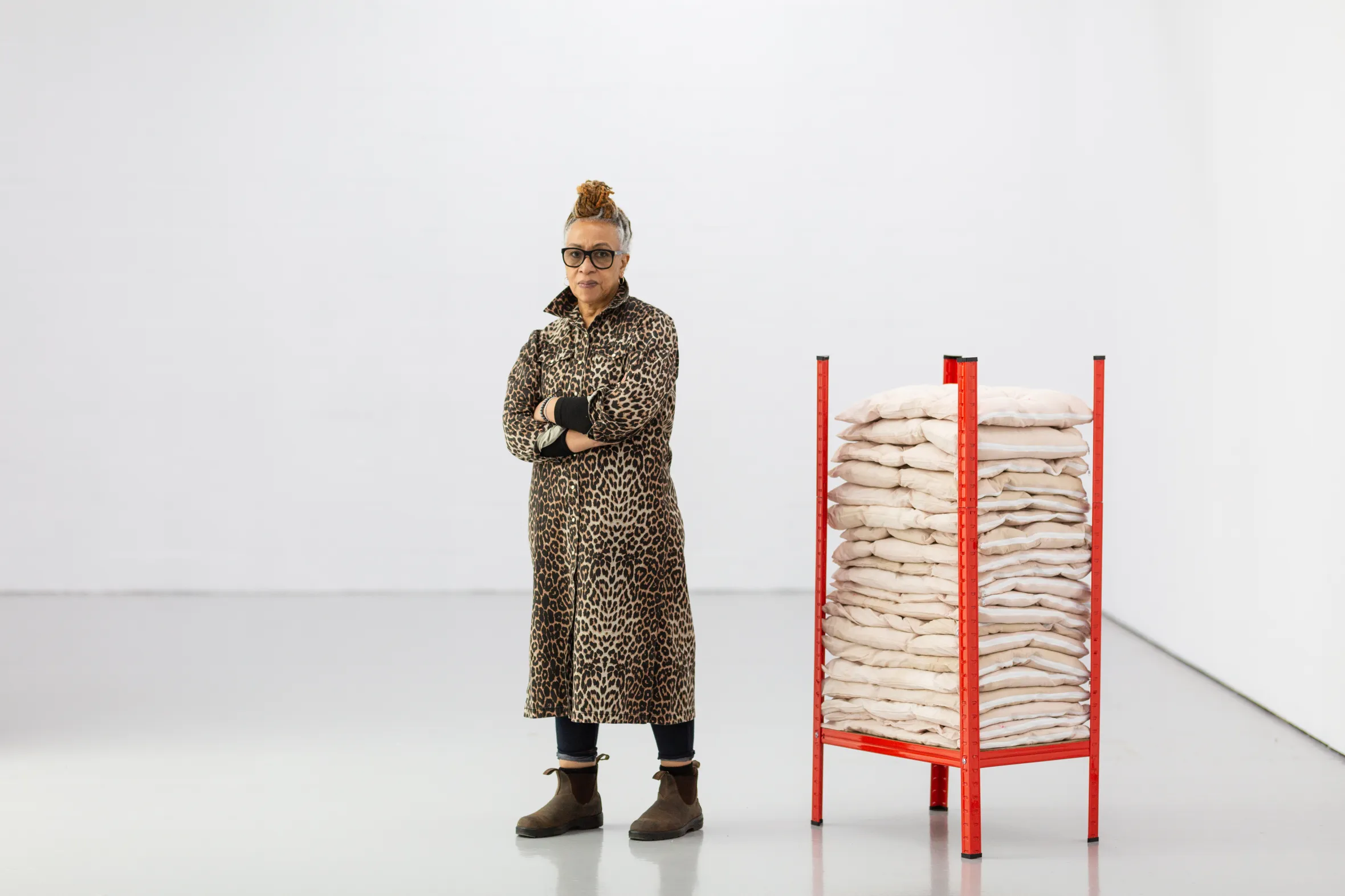 Veronica Ryan in a leopard print coat, standing with arms crossed next to a red shelving unit filled with light-colored sacks.