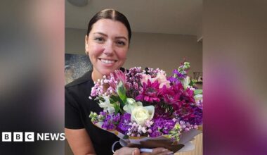 Natalie Dyson with brown hair in a pony tail and wearing gold earrings and black short sleeved top holding a bunch of white, purple and pink flowers hand tied towards the camera. She is standing in her kitchen. She is smiling.