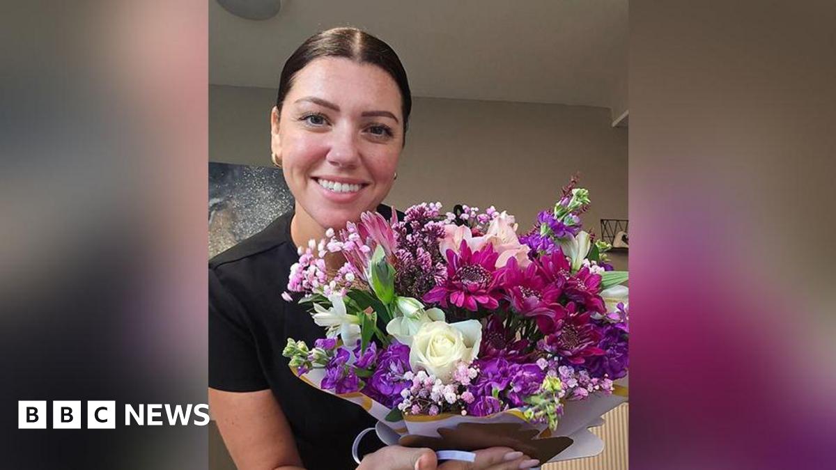 Natalie Dyson with brown hair in a pony tail and wearing gold earrings and black short sleeved top holding a bunch of white, purple and pink flowers hand tied towards the camera. She is standing in her kitchen. She is smiling.