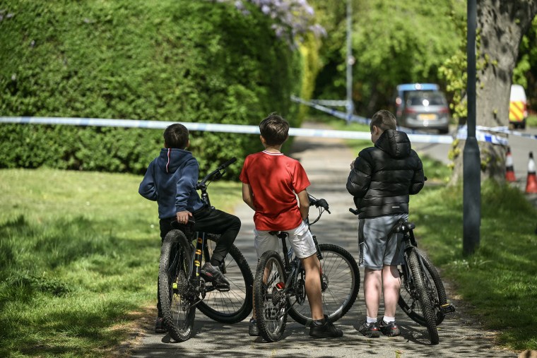 Three boys on bicycles face away from the camera and toward blue and white striped police tape. 