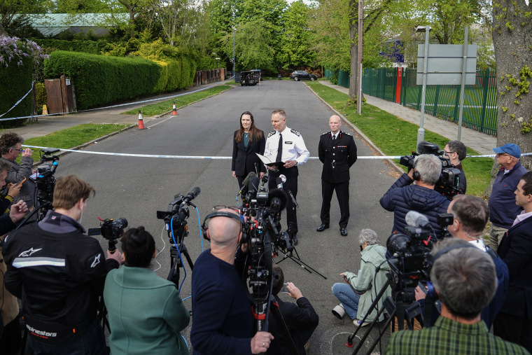 Three law enforcement officials stand in the middle of a street surrounded by reporters with cameras . 