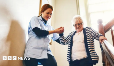 A care worker helping an elderly woman walk down a staircase. The carer, on the left, is wearing a light-blue jacket and dark trousers. She is smiling and holding the older woman's hand. The older woman has white hair, is wearing spectacles and has a look of concentration on her face as she takes the steps. She is holding the banister with her left hand.