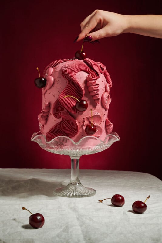 A tall, decorative pink cake topped with cherries sits on a glass stand against a dark red background. A hand is placing a cherry on top, with more cherries scattered on the white tablecloth below.