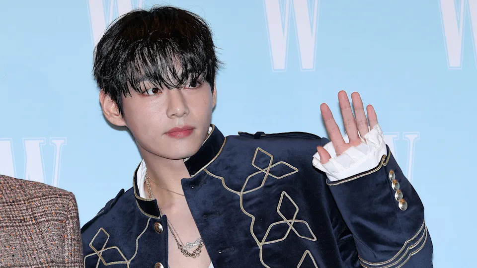 BTS singer V with dark hair wearing a navy blue jacket with gold embroidery, waving against a light blue background.