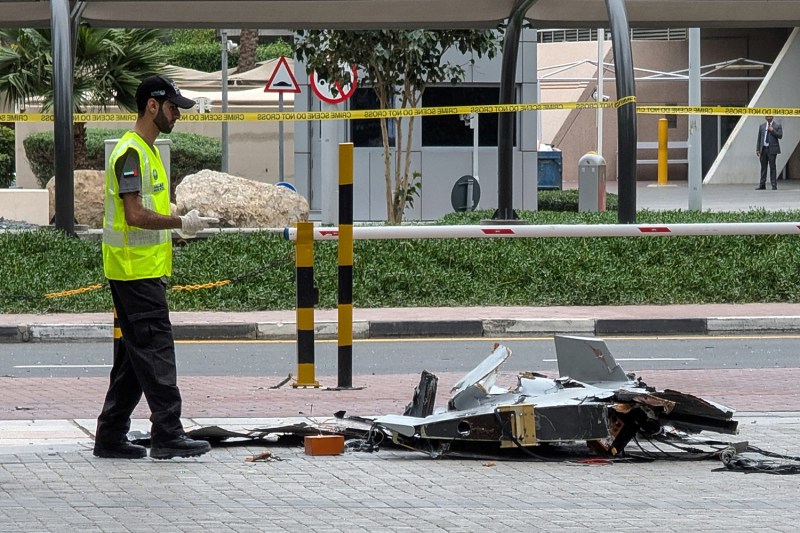 A man in a neon vest looks down at a crashed drone on the sidewalk.
