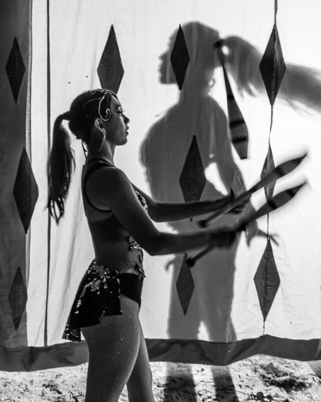 A woman in costume juggles clubs in front of a patterned curtain, casting a large shadow behind her. The scene is in black and white.