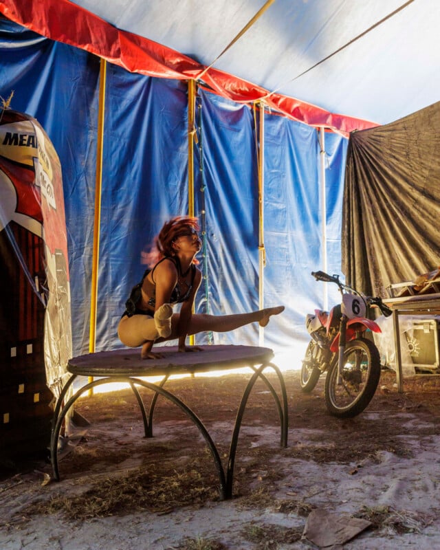 A contortionist performs on a round table inside a circus tent, with a motorcycle and equipment in the background. Blue and red fabric walls are visible, and sunlight streams in from the left.