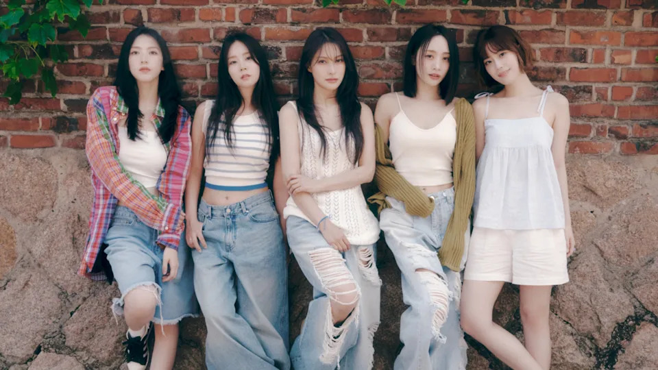 The members of KARA (circa 2024) pose casually against a brick wall, wearing casual summer outfits with denim and light tops.