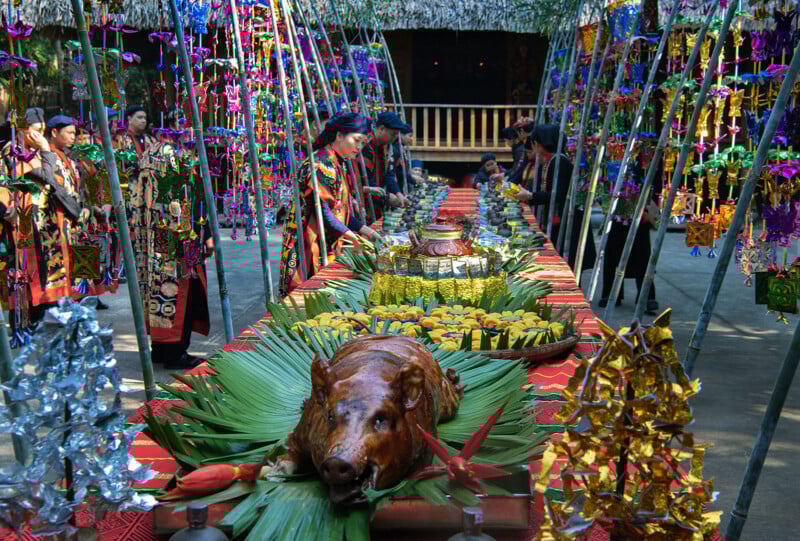 A festive, colorful table is set outdoors with a roast pig as the centerpiece, surrounded by traditional decorations, tropical leaves, and platters of food. People in vibrant cultural attire stand on either side.