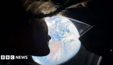 A woman wearing all black, with her arms crossed and smiling, in front of a white, blue and purple animated background full of shapes.