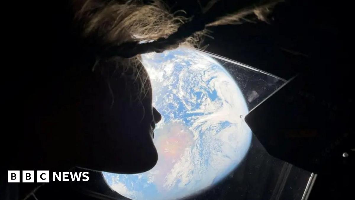 A woman wearing all black, with her arms crossed and smiling, in front of a white, blue and purple animated background full of shapes.