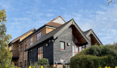 A landscape image of Farleigh Hospice's specialist inpatient unit in Chelmsford. It is a black wooden building in front of a brick building. It is surrounded by green bushes and grass and daffodils.