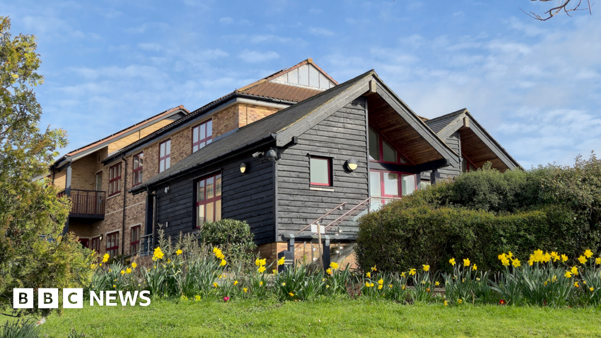A landscape image of Farleigh Hospice's specialist inpatient unit in Chelmsford. It is a black wooden building in front of a brick building. It is surrounded by green bushes and grass and daffodils.