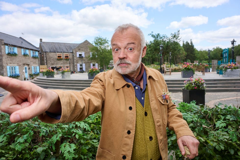 graham norton the neighbourhood man gesturing outdoors in a quaint town square with flowers and buildings