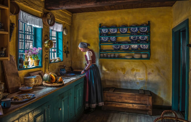 A woman in traditional clothing stands by a window in a rustic, old-fashioned kitchen with yellow walls, blue cabinets, flowers, and shelves holding blue and white dishes. Sunlight streams through the window.