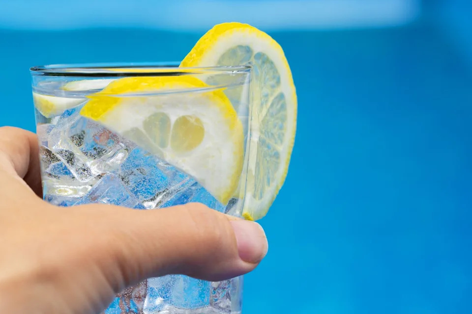 A hand holds a glass of iced water with lemon slices, set against a blurred blue background, suggesting a refreshing drink by the pool