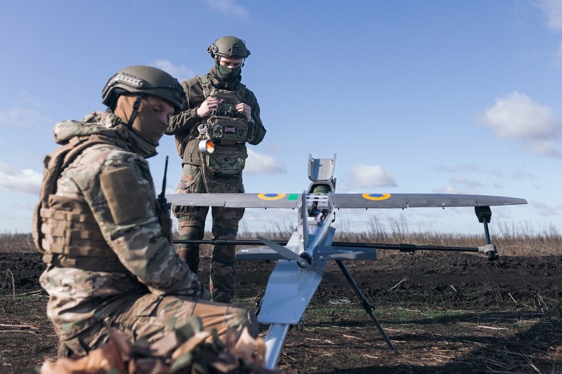 Two soldiers in uniform work with a drone, which is set up to be launched from the ground.