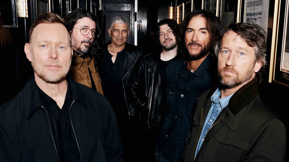 A promotional photograph of the Foo Fighters, showing the band lined up in a wood-panelled corridor, surrounded by picture frames