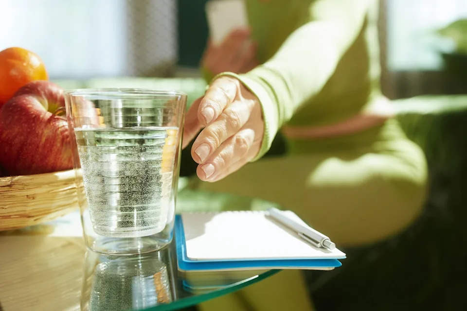 Person reaching for a glass of water on a table with a notepad, pen, and basket of apples nearby