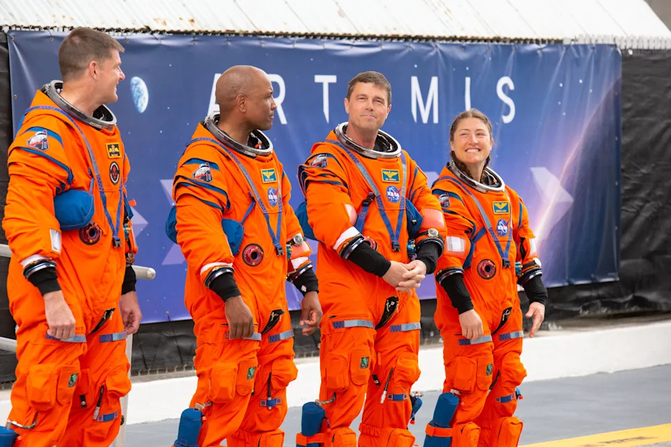 Four astronauts in orange space suits stand in front of an Artemis mission backdrop, preparing for a mission event