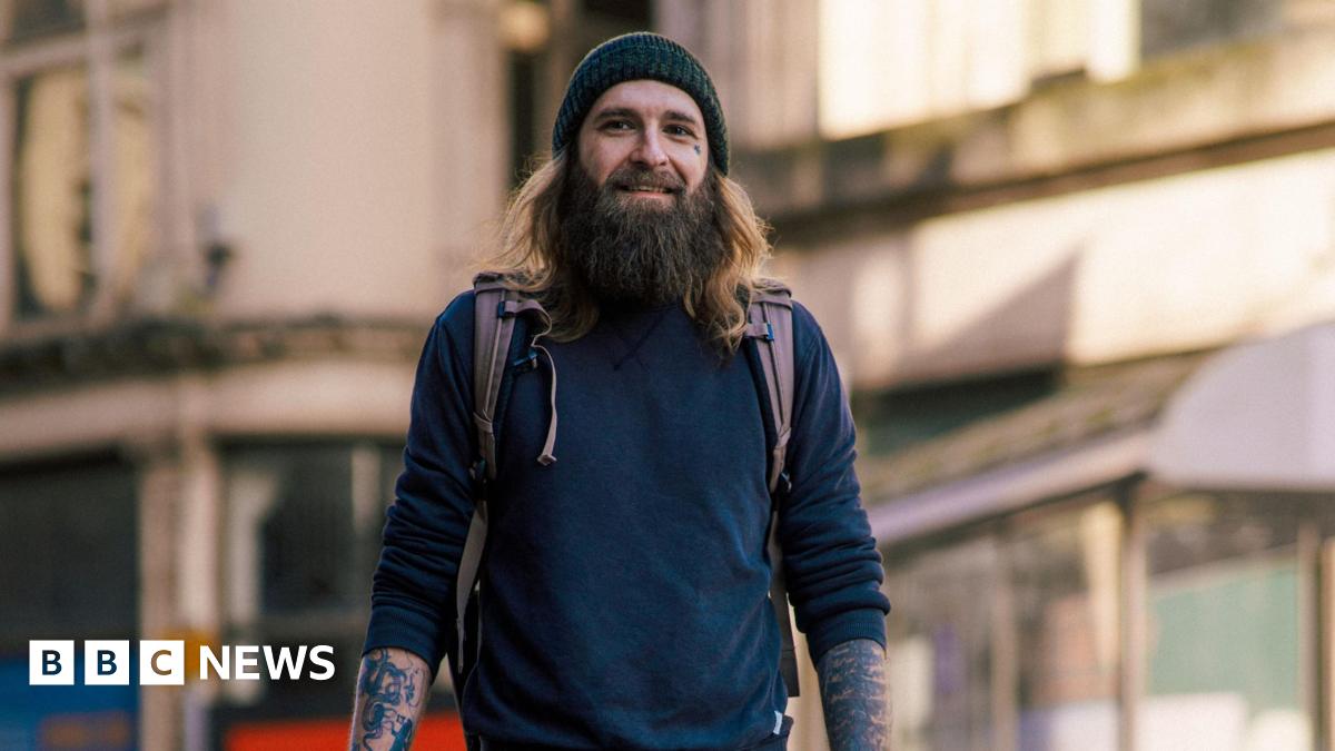 Tom Wilkins stood in Cardiff high street. He has a brown long beard, brown long hair and wears a knitted grey hat. Straps from his backpack can be seen on his shoulder, and he wears a jumper rolled up to his elbows