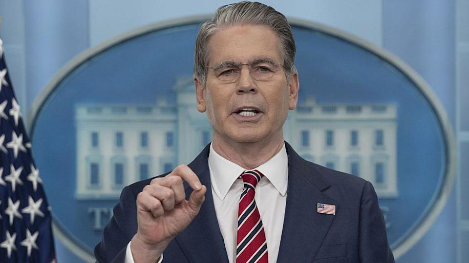 Face and shoulders of US Treasury Secretary Scott Bessent wearing a blue suit, white shirt, and red, white, and navy blue striped tie with a US flag lapel pin addressing the press, who are not in shot, in the White House press room. Behind him, there is US flag and and, mostly blocked by his face and body, a big image of the White House with the text 'The White House' beneath.