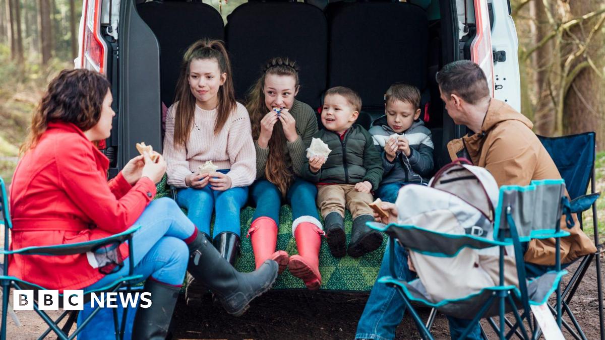 Children sitting together in the boot of a van in the woods eating packed lunch and two adults sitting on camping chairs outside.