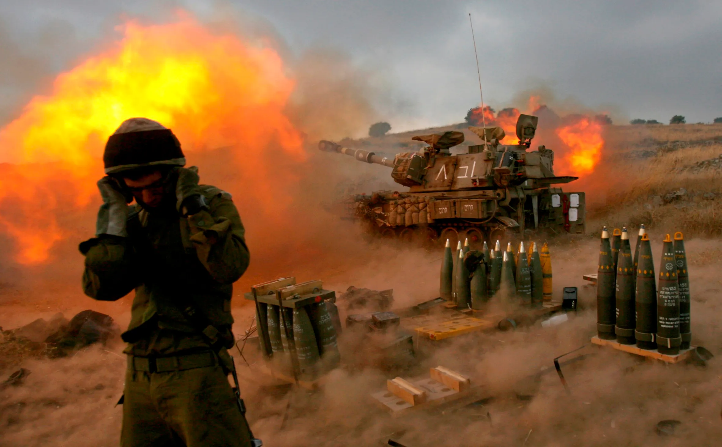An Israeli soldier standing near a mobile artillery unit as it fires a shell, with artillery shells on the ground.