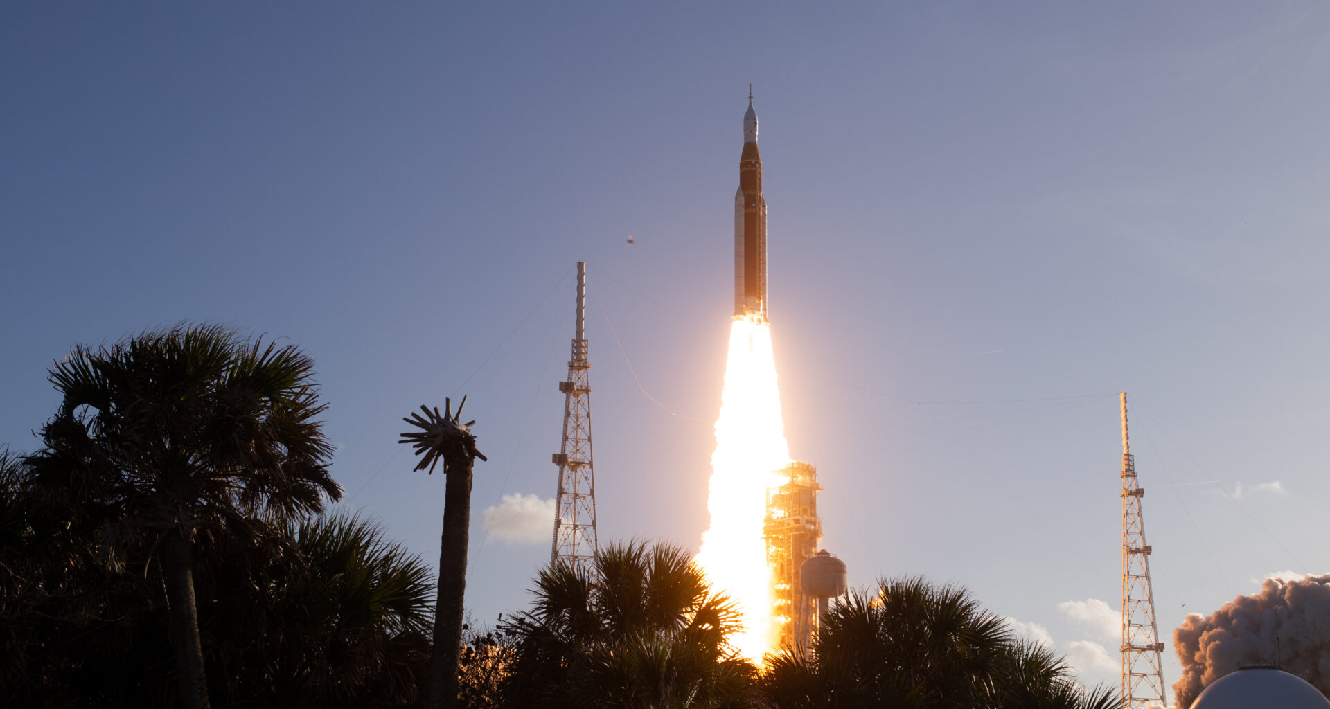 The Artemis II Moon rocket lifts off. There is a bright yellow-orange plume of fire underneath the rocket. The trees in the foreground appear in shadow.