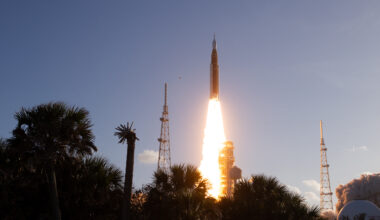 The Artemis II Moon rocket lifts off. There is a bright yellow-orange plume of fire underneath the rocket. The trees in the foreground appear in shadow.