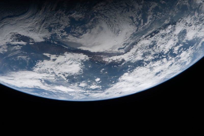 View of Earth from space showing clouds over land and ocean, with the curve of the planet visible against the darkness of space at the bottom of the image.