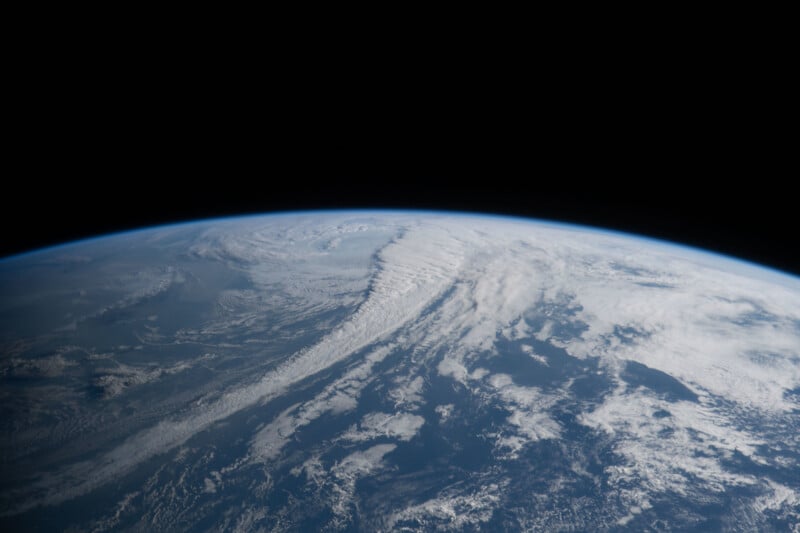 A view of Earth from space showing a curved horizon, swirling white clouds over blue oceans, and the blackness of outer space above.