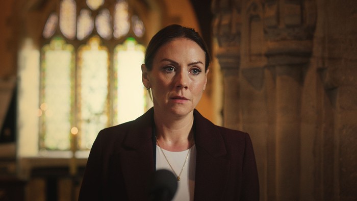 Stevie stands in a church in a scene from Casualty, wearing a white blouse and black jacket.