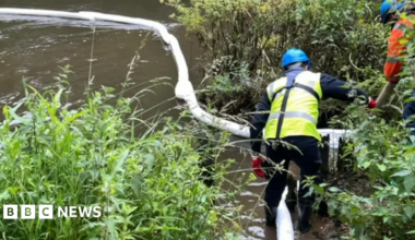 A helmeted worker stands in a river next to a floating bund which is being used to contain oil