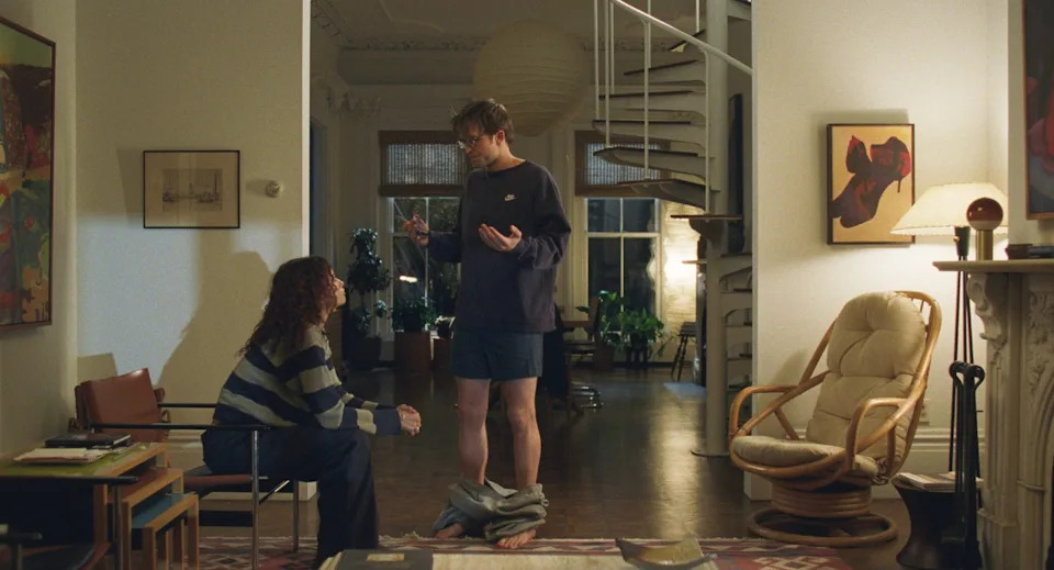 A person sitting on a couch talks to another standing in a spacious living room with a spiral staircase. The standing person appears to have just removed their pants