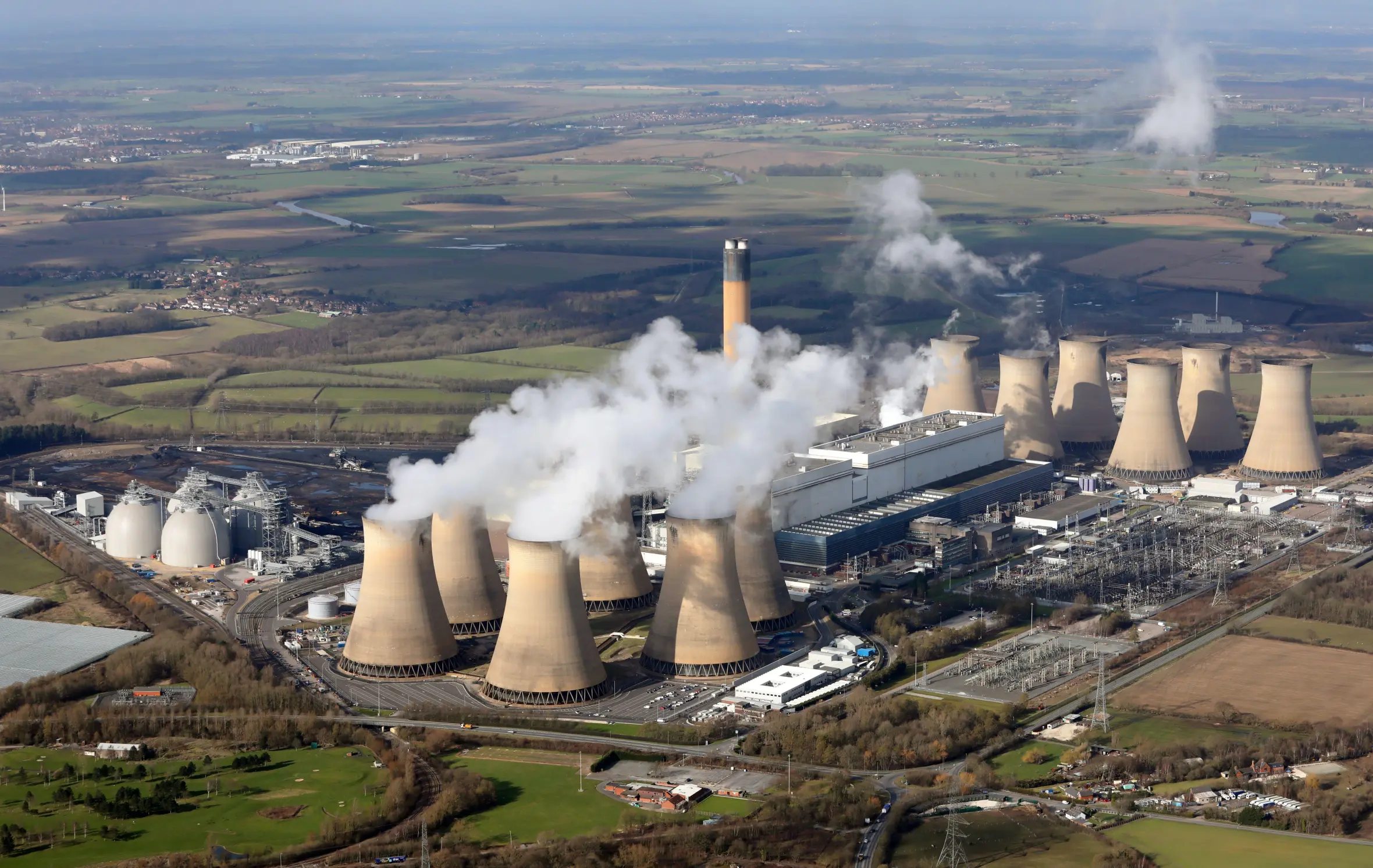 Aerial view of Drax power station with several cooling towers emitting steam, surrounded by fields and scattered buildings.
