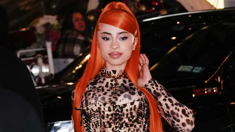 A woman with long orange hair and a leopard print outfit stands in front of a car, posing confidently.