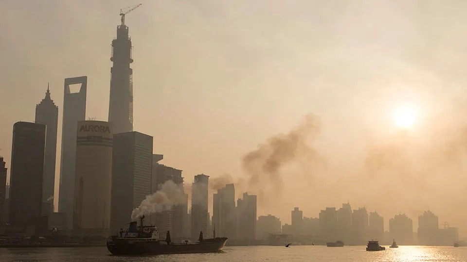 A smog-filled cityscape on the edge of a body of water is seen under a clouded over sun.